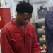 A U.S. Navy sailor named Rowe, wearing red coveralls, is working with equipment in what appears to be a shipboard workspace or maintenance area. He is focused on handling items in a foam-lined case or tray. The setting appears to be an interior compartment of a naval vessel.