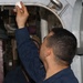 A U.S. Navy sailor is performing maintenance work near a watertight hatch or door aboard a naval vessel, applying what appears to be a cleaning or sealing material to the upper frame of the hatch. A second sailor is visible in the background. Both are wearing standard Navy blue coverall uniforms with U.S. flag and 'Don't Tread on Me' patches.