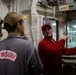Two individuals are inside what appears to be a naval vessel's interior compartment. One person wearing a gray jacket with 'INSURV' patch and a tan cap is being briefed by another person in a red jacket and red cap who is pointing toward a window or control panel. The setting contains electrical panels, cable bundles, and a labeled 'AFFF' (Aqueous Film-Forming Foam) fire suppression control panel.