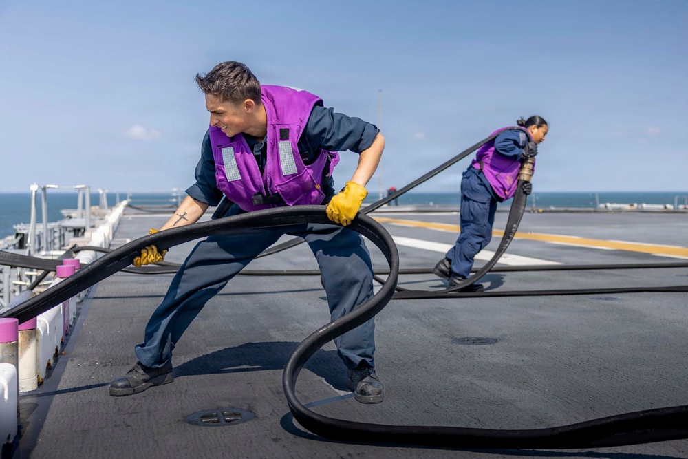 Two naval personnel are working on the flight deck of a military vessel, handling large black fuel or hydraulic hoses. Both individuals are wearing purple safety vests and yellow work gloves over navy blue coveralls, indicating they are part of a fueling or deck operations crew. The scene takes place on an open ocean with clear skies visible in the background.