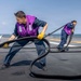 Two naval personnel are working on the flight deck of a military vessel, handling large black fuel or hydraulic hoses. Both individuals are wearing purple safety vests and yellow work gloves over navy blue coveralls, indicating they are part of a fueling or deck operations crew. The scene takes place on an open ocean with clear skies visible in the background.