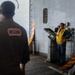 Two personnel are on the deck of a naval vessel, likely during an inspection or operational exercise. One individual wearing a jacket labeled 'INSURV' (Board of Inspection and Survey) stands with their back to the camera, while another person in a yellow safety vest and hearing protection operates a deck-mounted control station. The setting features characteristic naval ship markings including yellow and red hazard striping.