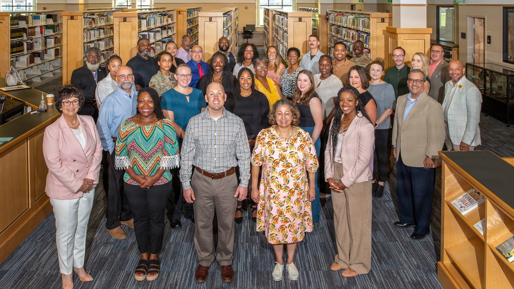 A large group of approximately 30 adults poses for a formal group photograph inside a library setting. The group is diverse in age, gender, and ethnicity, and appears to be dressed in business or business-casual attire, suggesting an organizational, institutional, or professional gathering. The setting features tall bookshelves lining both sides of the aisle behind the group.