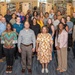 A large group of approximately 30 adults poses for a formal group photograph inside a library setting. The group is diverse in age, gender, and ethnicity, and appears to be dressed in business or business-casual attire, suggesting an organizational, institutional, or professional gathering. The setting features tall bookshelves lining both sides of the aisle behind the group.