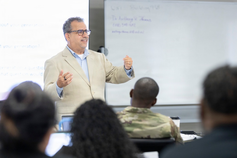 A middle-aged man in a beige blazer and glasses is presenting or lecturing to a seated audience in what appears to be a classroom or conference room setting. Behind him are two whiteboards with handwritten text, one of which appears to contain contact information. At least one audience member is wearing military camouflage attire.