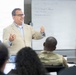 A middle-aged man in a beige blazer and glasses is presenting or lecturing to a seated audience in what appears to be a classroom or conference room setting. Behind him are two whiteboards with handwritten text, one of which appears to contain contact information. At least one audience member is wearing military camouflage attire.