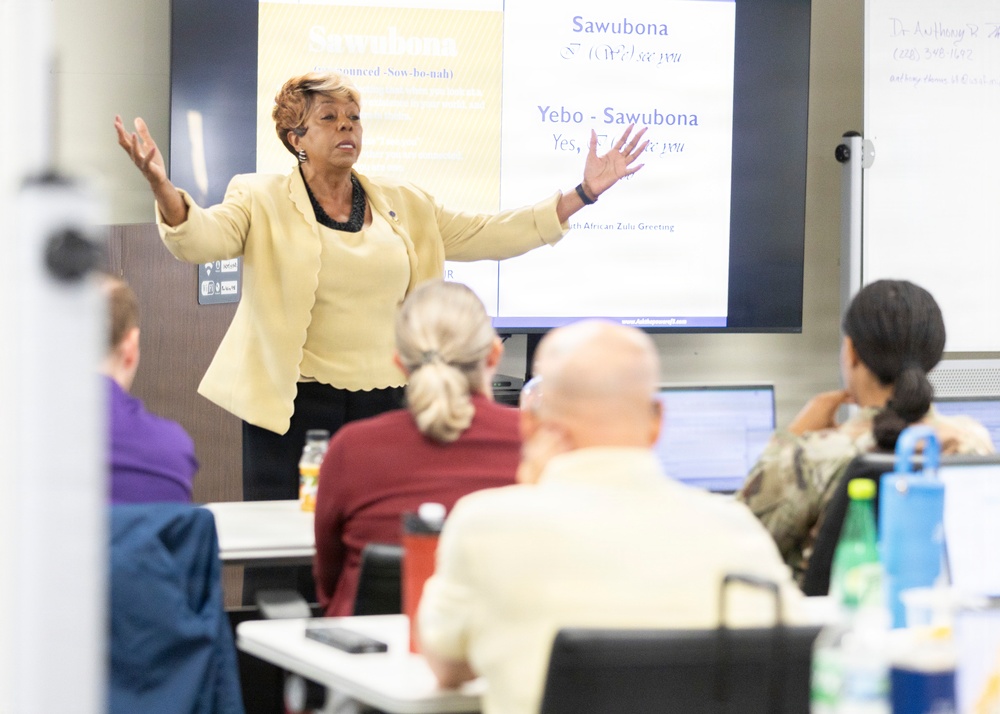 A woman in a yellow blazer is presenting to a classroom audience, gesturing expressively with both arms raised. The presentation slide behind her displays Zulu language greetings, specifically 'Sawubona' and 'Yebo - Sawubona,' identified as South African Zulu greetings. Several attendees are seated at desks facing the presenter, with laptops and personal items visible.