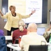 A woman in a yellow blazer is presenting to a classroom audience, gesturing expressively with both arms raised. The presentation slide behind her displays Zulu language greetings, specifically 'Sawubona' and 'Yebo - Sawubona,' identified as South African Zulu greetings. Several attendees are seated at desks facing the presenter, with laptops and personal items visible.