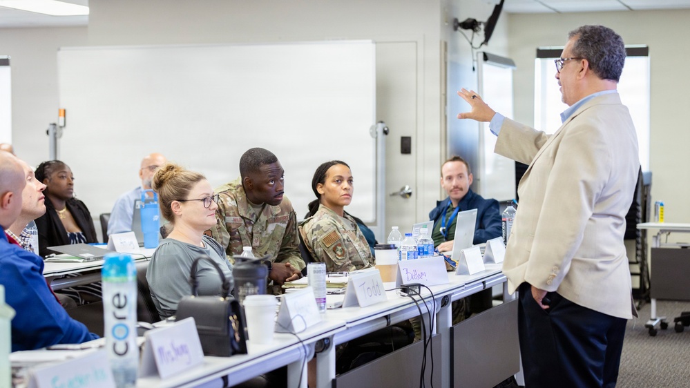 A professional training or briefing session is taking place in a modern classroom or conference room setting. A civilian instructor or presenter in a beige blazer is addressing a mixed audience that includes both military personnel in camouflage uniforms and civilians seated at tables. Name placards are visible on the tables, suggesting a formal structured learning environment.