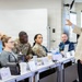 A professional training or briefing session is taking place in a modern classroom or conference room setting. A civilian instructor or presenter in a beige blazer is addressing a mixed audience that includes both military personnel in camouflage uniforms and civilians seated at tables. Name placards are visible on the tables, suggesting a formal structured learning environment.
