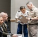 A group of U.S. military personnel from multiple branches are gathered in what appears to be a conference or briefing room. Two service members in the foreground — one in U.S. Army dress whites with blue trousers and one in U.S. Navy/Marine Corps tan service uniform — are reviewing documents on a clipboard. Several other uniformed personnel are seated in the background.