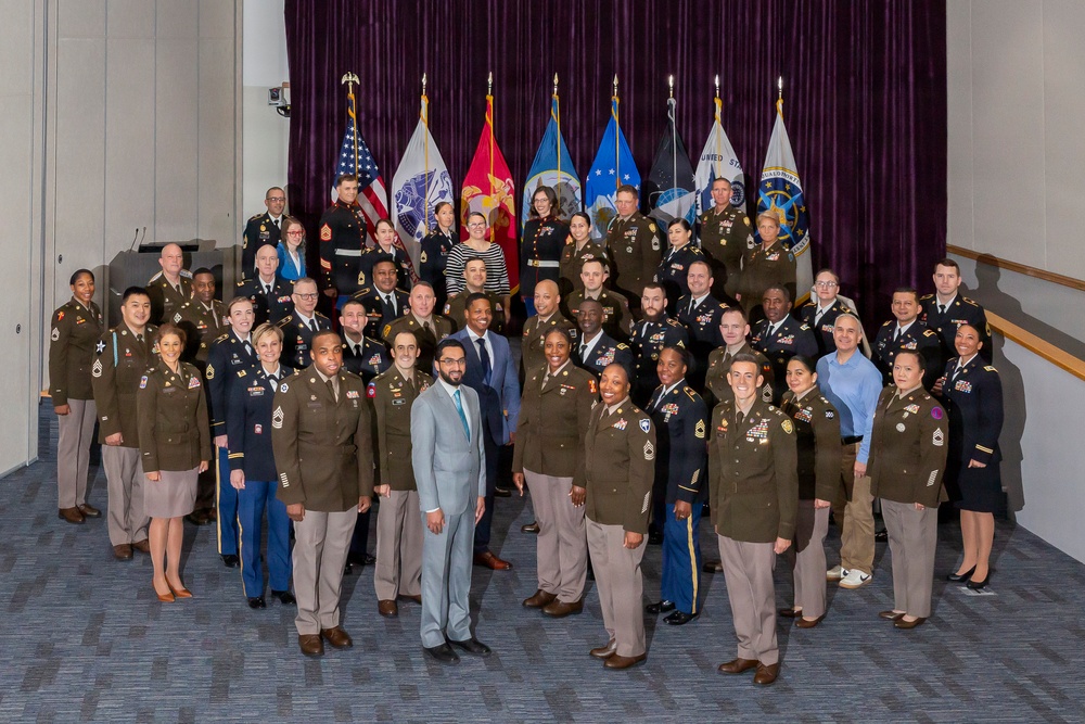 A formal group photograph of approximately 50 individuals, predominantly U.S. Army personnel in olive/brown service uniforms, along with a few civilians and one Marine Corps member. The group is posed in tiered rows in front of multiple military branch flags and a deep purple curtain backdrop. This appears to be an official military ceremony, graduation, or organizational group photo.