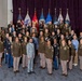 A formal group photograph of approximately 50 individuals, predominantly U.S. Army personnel in olive/brown service uniforms, along with a few civilians and one Marine Corps member. The group is posed in tiered rows in front of multiple military branch flags and a deep purple curtain backdrop. This appears to be an official military ceremony, graduation, or organizational group photo.