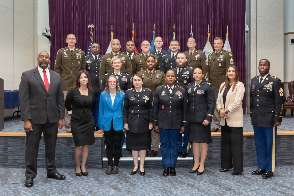 A formal group photograph of approximately 20 individuals, a mix of U.S. Army military personnel in uniform and civilians in business attire, posed in three rows in what appears to be an official government or military facility. Multiple flags are visible in the background against a purple curtain backdrop, suggesting a ceremonial or graduation event. The group includes both men and women of diverse backgrounds.
