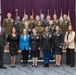 A formal group photograph of approximately 20 individuals, a mix of U.S. Army military personnel in uniform and civilians in business attire, posed in three rows in what appears to be an official government or military facility. Multiple flags are visible in the background against a purple curtain backdrop, suggesting a ceremonial or graduation event. The group includes both men and women of diverse backgrounds.