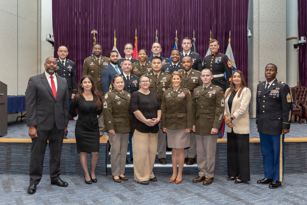 A formal group photograph of approximately 20 individuals, the majority wearing U.S. military uniforms (primarily U.S. Army dress uniforms with one U.S. Marine Corps dress uniform visible), along with several civilians in business attire. The group is posed in two rows inside what appears to be a formal auditorium or conference room with purple curtain backdrop and military flags. This appears to be an official military ceremony, graduation, or recognition event.