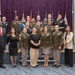 A formal group photograph of approximately 20 individuals, the majority wearing U.S. military uniforms (primarily U.S. Army dress uniforms with one U.S. Marine Corps dress uniform visible), along with several civilians in business attire. The group is posed in two rows inside what appears to be a formal auditorium or conference room with purple curtain backdrop and military flags. This appears to be an official military ceremony, graduation, or recognition event.