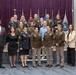 A formal group photograph of approximately 18-20 individuals, the majority wearing U.S. Army service uniforms (Army Service Uniform in olive/green), with at least one individual in U.S. Marine Corps dress uniform. The group is posed in front of multiple military unit and organizational flags against a burgundy/purple curtain backdrop, suggesting an official military ceremony or graduation event. Several civilians in business attire are also present among the group.