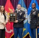 A military award ceremony at the Defense Equal Opportunity Management Institute (DEOMI), where a soldier in Army dress uniform is receiving a Commandant's Award plaque. He is flanked by a civilian woman in a white blazer on his left and a senior non-commissioned officer (Sergeant Major) in Army dress uniform on his right. Multiple military branch flags are displayed in the background.