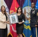 Three individuals pose for a formal photograph in front of multiple military service flags. Two civilian women jointly hold a framed award or certificate bearing the text 'DEOMI,' while a U.S. Army Sergeant Major in dress uniform stands to the right. The setting appears to be an official military ceremony or recognition event.