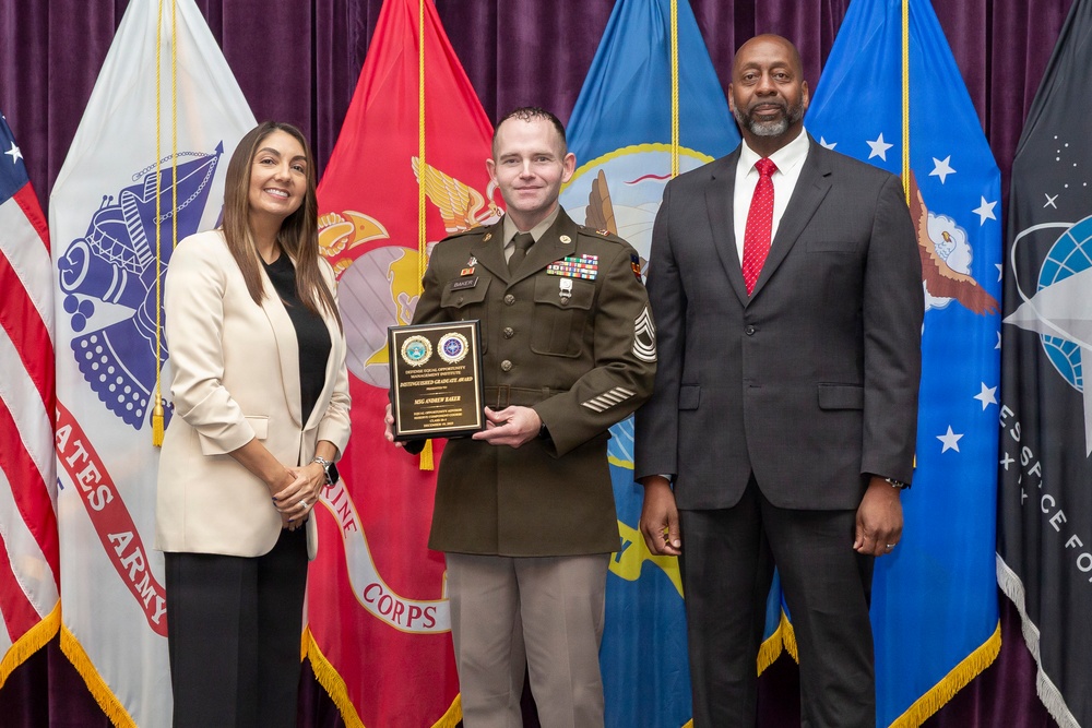 A formal military award ceremony featuring three individuals posing for a photo. A U.S. Army soldier in dress uniform holds a wooden plaque award, flanked by a woman in a white blazer and a man in a dark suit with a red tie. Multiple military branch flags are displayed in the background.