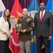 A formal military award ceremony featuring three individuals posing for a photo. A U.S. Army soldier in dress uniform holds a wooden plaque award, flanked by a woman in a white blazer and a man in a dark suit with a red tie. Multiple military branch flags are displayed in the background.