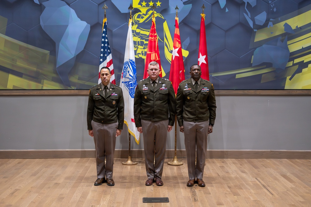 Three U.S. Army officers in dress green uniforms are posed formally in front of a display of military flags, including the American flag and what appear to be general officer flags with stars. The setting appears to be an official military headquarters or command facility, featuring a modern decorative wall mural with geometric blue and yellow patterns. This appears to be an official military portrait or ceremony photograph.