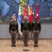 Three U.S. Army officers in dress green uniforms are posed formally in front of a display of military flags, including the American flag and what appear to be general officer flags with stars. The setting appears to be an official military headquarters or command facility, featuring a modern decorative wall mural with geometric blue and yellow patterns. This appears to be an official military portrait or ceremony photograph.