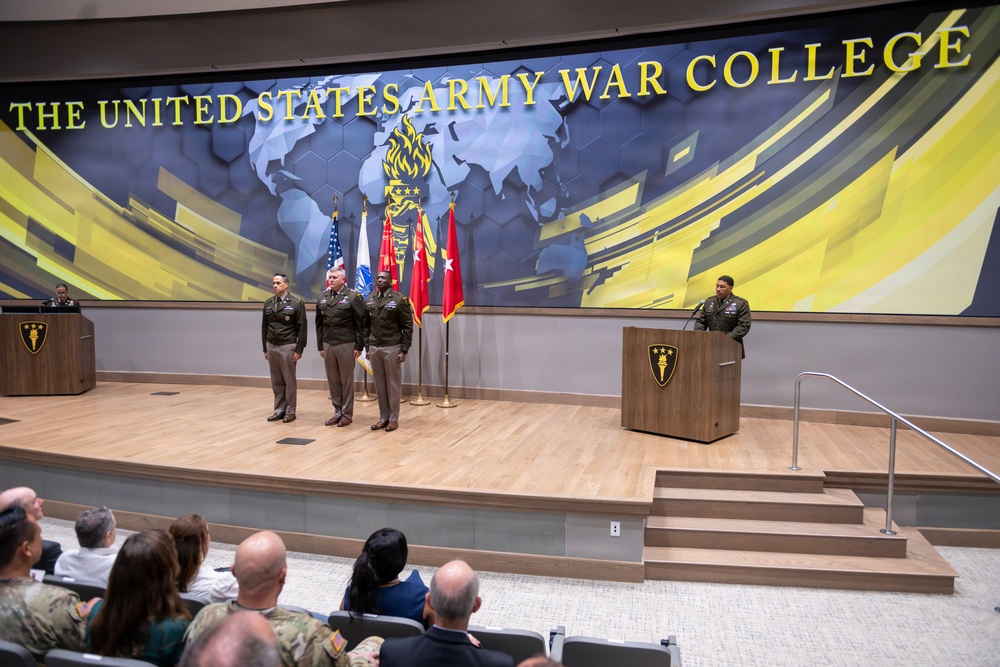 A formal military ceremony is taking place at The United States Army War College, visible from the large backdrop signage. Several uniformed military officers are standing on a raised stage, while one officer addresses the audience from a podium. An audience of military and civilian personnel is seated in the foreground observing the event.