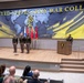 A formal military ceremony is taking place at The United States Army War College, visible from the large backdrop signage. Several uniformed military officers are standing on a raised stage, while one officer addresses the audience from a podium. An audience of military and civilian personnel is seated in the foreground observing the event.