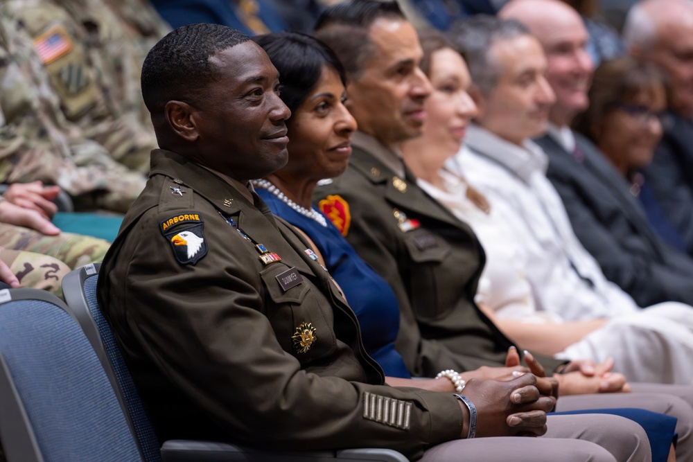 A group of military personnel and civilians are seated in rows, likely attending a formal ceremony or briefing. The foreground features a U.S. Army soldier wearing the 101st Airborne Division patch and service uniform with visible ribbons and insignia. The atmosphere appears formal and attentive.