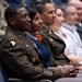A group of military personnel and civilians are seated in rows, likely attending a formal ceremony or briefing. The foreground features a U.S. Army soldier wearing the 101st Airborne Division patch and service uniform with visible ribbons and insignia. The atmosphere appears formal and attentive.