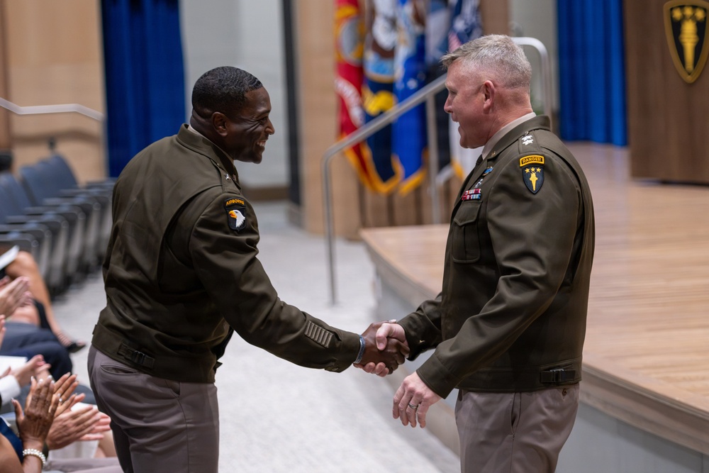 Two U.S. Army officers in Army Green Service Uniforms are shaking hands during what appears to be a formal military ceremony, likely a change of command or award presentation. Both officers display general officer rank insignia and unit patches, including a 101st Airborne Division patch on the left officer and a Ranger tab on the right officer. An audience is visible applauding in the background.