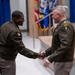 Two U.S. Army officers in Army Green Service Uniforms are shaking hands during what appears to be a formal military ceremony, likely a change of command or award presentation. Both officers display general officer rank insignia and unit patches, including a 101st Airborne Division patch on the left officer and a Ranger tab on the right officer. An audience is visible applauding in the background.