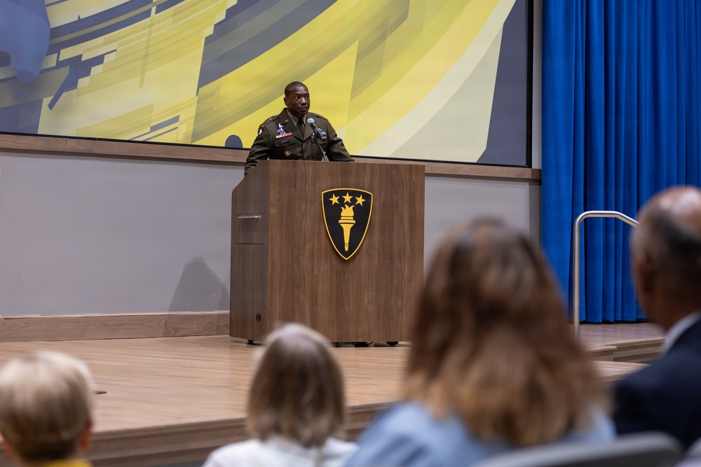 A U.S. Army officer in olive green service uniform is delivering a speech at a wooden podium on a stage. The podium displays the insignia of U.S. Army Europe and Africa (shield with four stars and a torch). An audience is seated in the foreground watching the presentation.