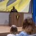 A U.S. Army officer in olive green service uniform is delivering a speech at a wooden podium on a stage. The podium displays the insignia of U.S. Army Europe and Africa (shield with four stars and a torch). An audience is seated in the foreground watching the presentation.
