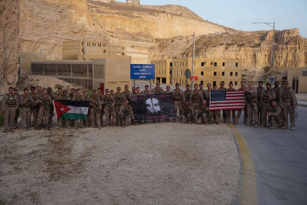 A large group of military personnel in desert camouflage uniforms pose for a group photograph at what appears to be a military installation or training facility. They are holding three flags: the Jordanian flag on the left, a unit/unofficial banner in the center, and the United States flag on the right. A blue sign in the background reads 'HIGH QUALITY AREA' in both Arabic and English, suggesting a location in Jordan.