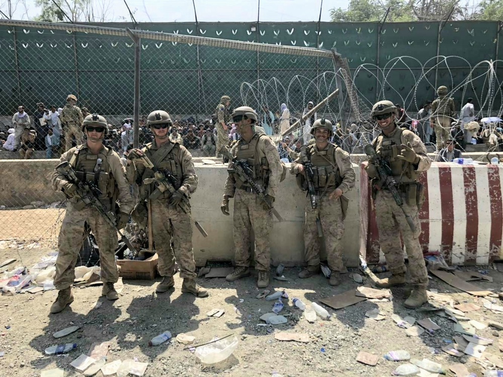Five armed military personnel in desert camouflage uniforms pose in front of a concrete barrier and chain-link fence topped with concertina/razor wire. Behind the fence, a large crowd of civilians is visible. The ground in the foreground is littered with debris including plastic bottles, cardboard, and discarded items.
