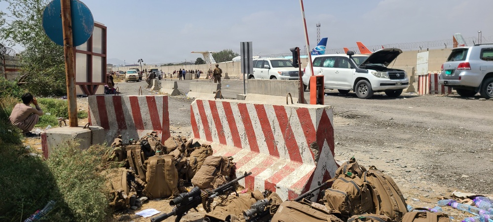 The image shows a security checkpoint at what appears to be an airport perimeter, with red-and-white striped concrete barriers in the foreground. Military tactical gear, backpacks, and firearms are piled on the ground near the barriers. Commercial aircraft and vehicles are visible in the background beyond the checkpoint gate.