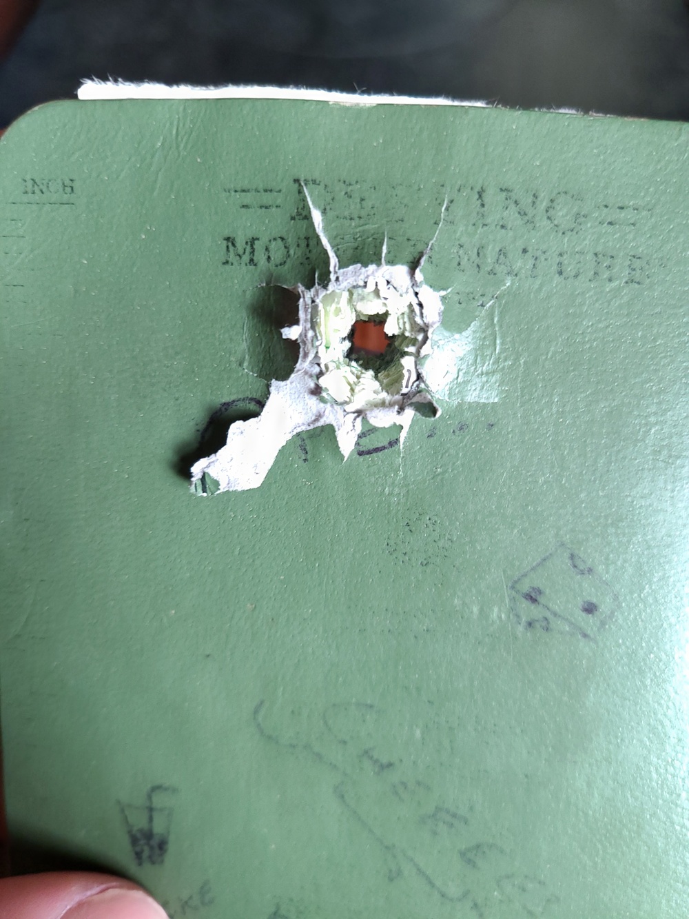 The image shows a green notebook or journal with a jagged hole punched or torn through its cover. The hole has exposed the white inner layers of the cover material and reveals a reddish-brown surface beneath. Partial text is visible on the cover reading what appears to be 'DEFIANCE MOTHER NATURE' along with handwritten markings.
