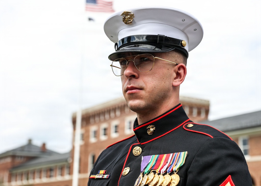 A United States Marine Corps service member stands at attention in dress blue uniform during what appears to be a formal military ceremony or parade. The Marine is wearing the distinctive USMC dress blues with red piping, white cover (hat), and multiple medals and ribbons on his chest. An American flag is visible in the background along with a brick building consistent with a military installation.