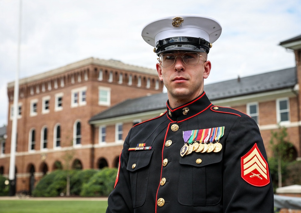 A United States Marine Corps Sergeant stands at attention in dress blue uniform at what appears to be a formal military installation. The Marine displays multiple medals and ribbons on his left chest, indicating significant service accomplishments. The background features a brick building consistent with a military base or government facility.