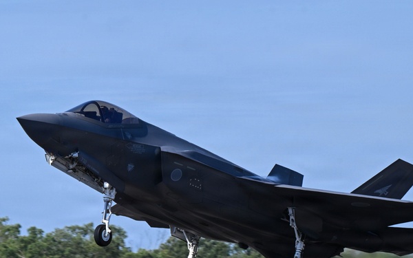 A dark-colored fifth-generation stealth fighter jet, consistent with an F-35 Lightning II, is captured during takeoff or low-altitude flight with landing gear still extended. The aircraft is photographed from a slightly elevated angle against a clear blue sky with a tree line and airfield perimeter fence visible in the background. The image is sharp and well-composed, suggesting professional aviation photography.