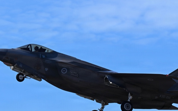 A dark-colored F-35 Lightning II stealth fighter jet is captured in flight with landing gear fully deployed, suggesting an approach to landing. The aircraft bears visible markings consistent with the Royal Australian Air Force (RAAF), including a roundel insignia and the serial number 'A35-016' on the fuselage. The image is taken against a clear blue sky with light cloud cover.