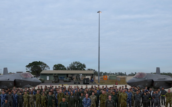 A large group of military personnel from multiple nations pose for a formal group photograph on an airfield tarmac in front of two F-35 Lightning II stealth fighter jets. Personnel are wearing a variety of uniforms including olive/tan camouflage, blue camouflage, and dark flight suits, suggesting a multinational military exercise or joint operation. Three individuals stand prominently in the foreground, likely senior officers or commanders.