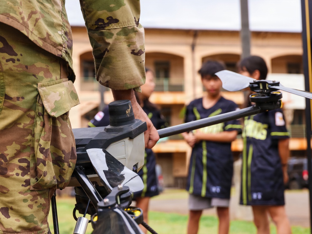 A military personnel in camouflage uniform holds a drone or unmanned aerial vehicle (UAV) while several children in matching dark athletic uniforms observe in the background. The setting appears to be an outdoor courtyard or parade ground near a multi-story building. This appears to be an educational or outreach demonstration event.