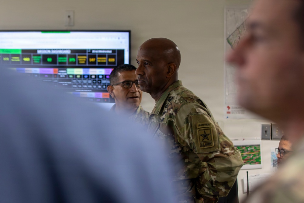 A U.S. Army soldier wearing OCP (Operational Camouflage Pattern) uniform with a Ranger tab and U.S. Army patch is engaged in a briefing or discussion in what appears to be a military operations center or command room. A large monitor displaying a 'Mission Dashboard' with color-coded status indicators is visible in the background. Several other individuals, both military and possibly civilian, are present in the room.