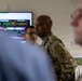 A U.S. Army soldier wearing OCP (Operational Camouflage Pattern) uniform with a Ranger tab and U.S. Army patch is engaged in a briefing or discussion in what appears to be a military operations center or command room. A large monitor displaying a 'Mission Dashboard' with color-coded status indicators is visible in the background. Several other individuals, both military and possibly civilian, are present in the room.