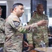 A group of U.S. Army soldiers in OCP (Operational Camouflage Pattern) uniforms are gathered in what appears to be a military operations or briefing room. The central figure is gesturing and appears to be presenting or explaining something at a table. The setting suggests a command post, planning session, or unit briefing environment.