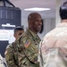 A group of U.S. Army soldiers in Operational Camouflage Pattern (OCP) uniforms are gathered in what appears to be an indoor military facility. The central figure, a bald male soldier, is smiling and appears to be engaged in conversation. He wears a U.S. flag patch and an Airborne tab with a unit patch on his left sleeve.